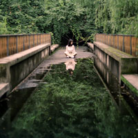 A woman sits at the end of a bridge with a reflecting pool of water.