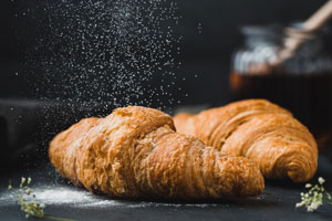 Two flaky croissants are being sprinkled with powdered sugar. 