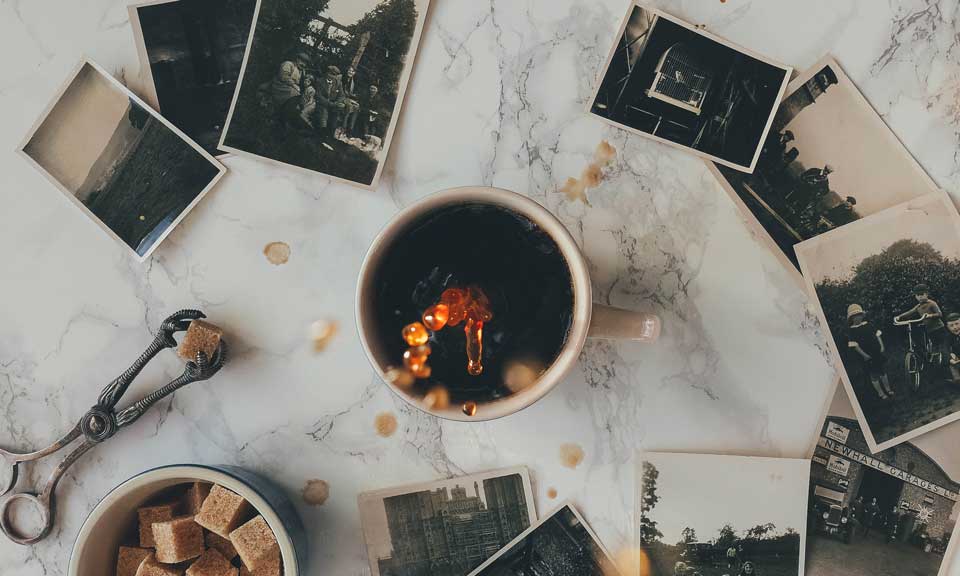 An aerial view of a cup of black coffee surrounded by old-style photographs and antique serveware. 