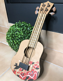 A cardboard Ukulele with decorative red flowers on the body leans against a small fake plant
