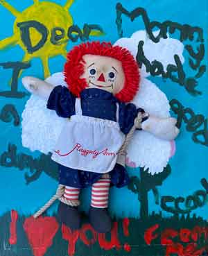 A Raggedy Ann doll attached to a canvas with a childlike paintinf of wings, the sun, and grass behind her along with text of 'Dear Mommy I hade a good day at school I love you From Me' in childlike writing