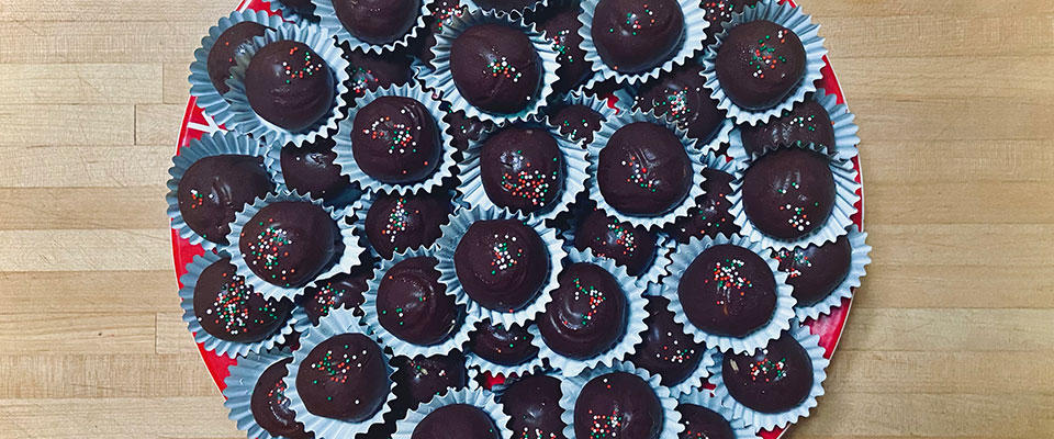 On a large tray are many stacked chocolate covered peanut butter balls. Each peanut butter ball is sitting in their own small muffin tin and topped with sprinkles