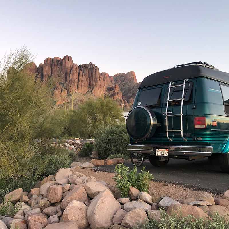 A green chevy g20 backed up to a campspot amongst rocks with a layered desert mountain in the background