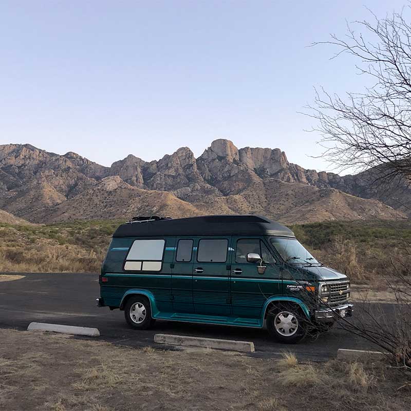 A checy g20 van sits parked with a desert mountain in the background