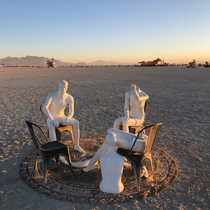 Three manequins positioned in the middle of the desert sitting on chairs and a rug