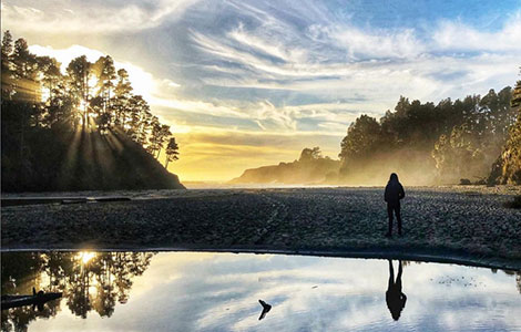 Girl Standing on the beach at sunset with reflection in the water looking out at the ocean with sun rays on it