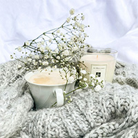 still photo of a cup of coffee, a candle, and flowers, grouped together on top of a crochet blanket.