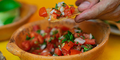 A person holding a chip with salsa over a bowl with more salsa.