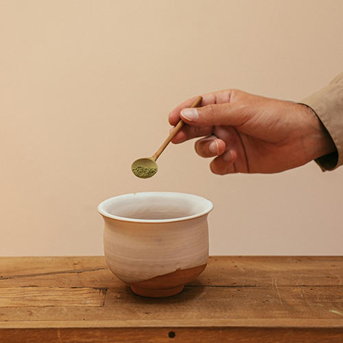 Hand holding a wooden spoon full of matcha powder over a clay bowl.