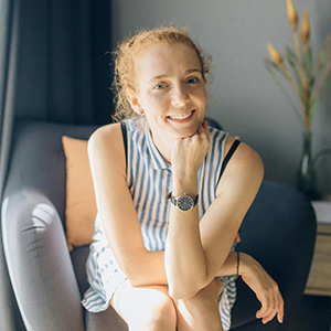 A smiling woman with red curly hair, wearing a blue and white shirt, and wearing a watch.