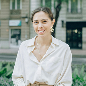 A smiling women with brown hair, wearing a white blouse, and gold earrings.