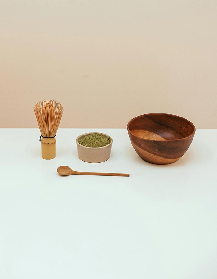 A white table with a wooden whisk, spoon, bowl, and container full of matcha on top.
