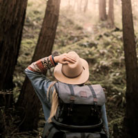 A women with a hat and backpack looks up into the trees while hiking.