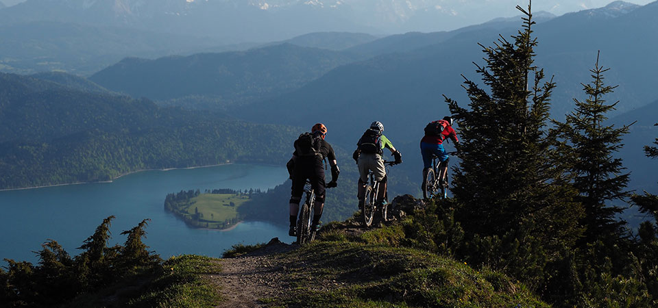 three bike riders on a trail in forested mountains overlooking a lake