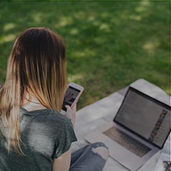 Woman sitting on grass looking at her phone with her computer open.