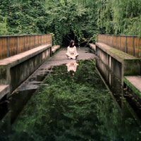 A woman sits at the end of a bridge with a reflecting pool of waterin front of her