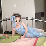 Image of child dressed in blue dress laying on bed with sunglasses on