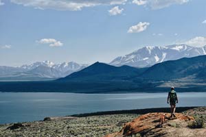 A young man and a dog wandering along the edge of a cliff overlooking a lake and a mountain range.