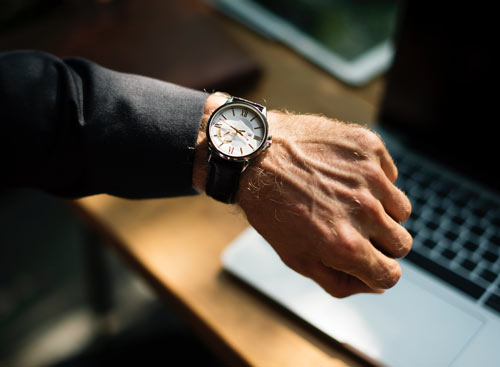 arm extended to the front of the camera displaying accessory, watch, background is a desk and computer blurred out.